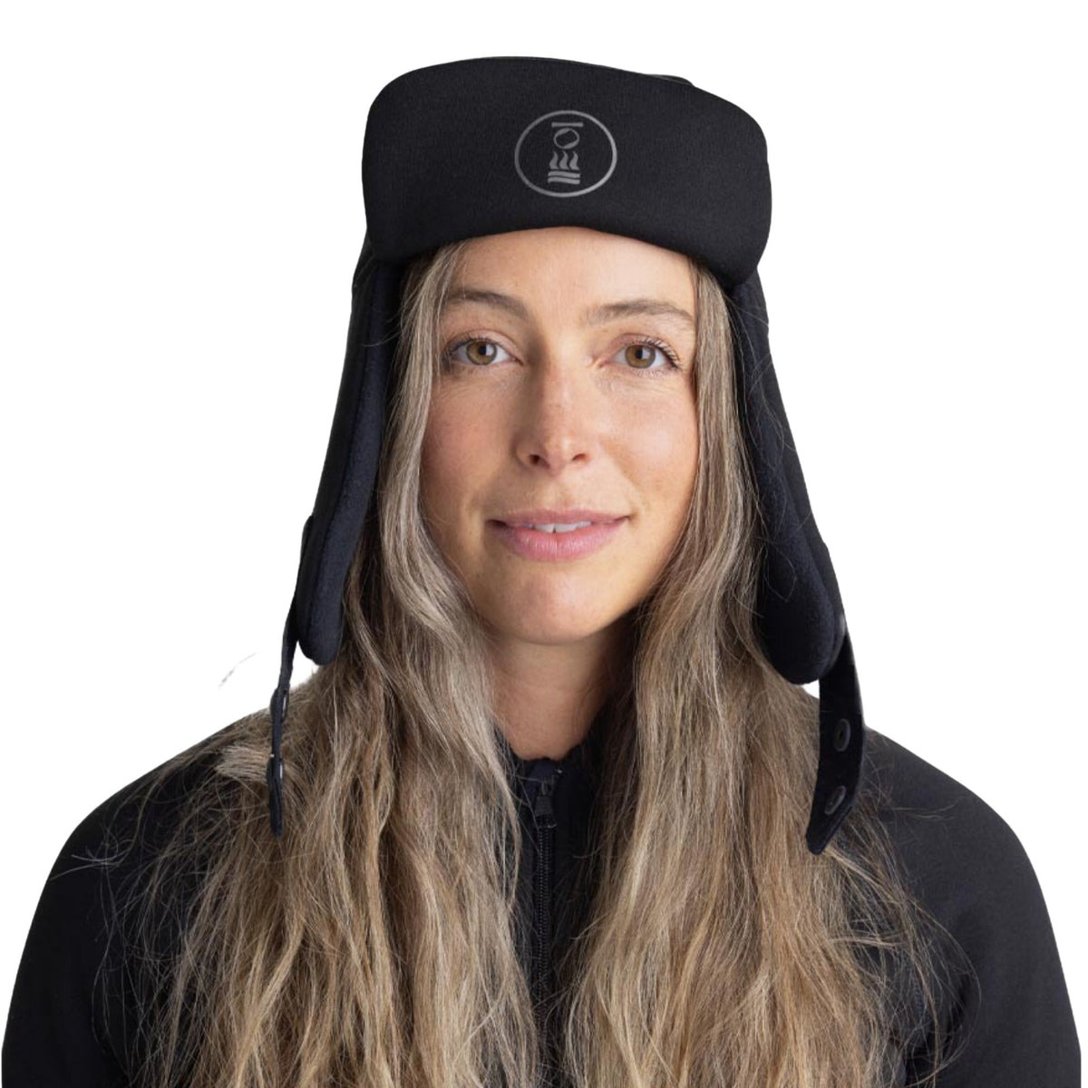 A woman with long blonde hair wearing a black insulated winter trapper hat with ear flaps and a small white circular logo on the front, set against a plain white background.