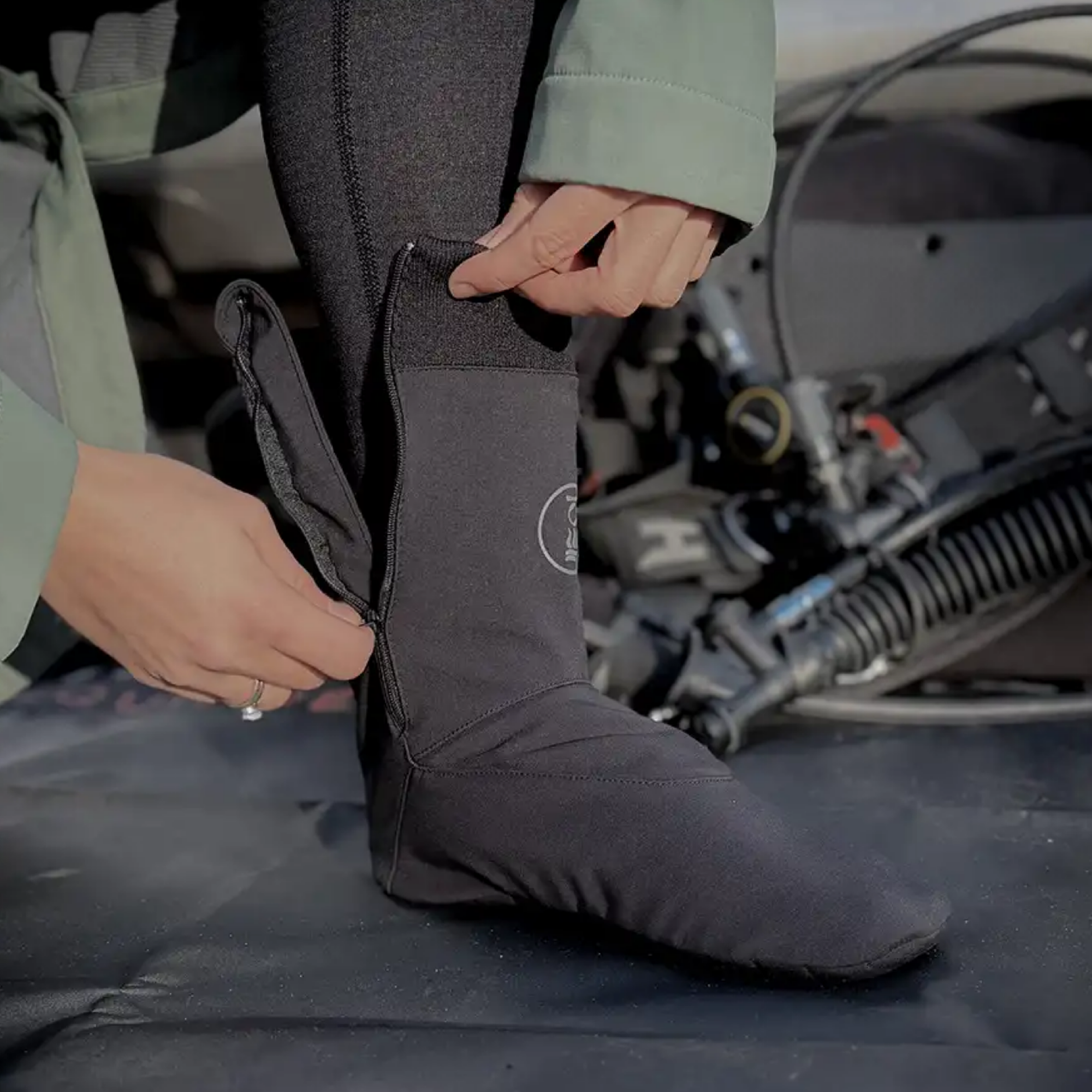 A person wearing a sock and pulling the zipper open with both hands, set against a blurred background of dive gear.