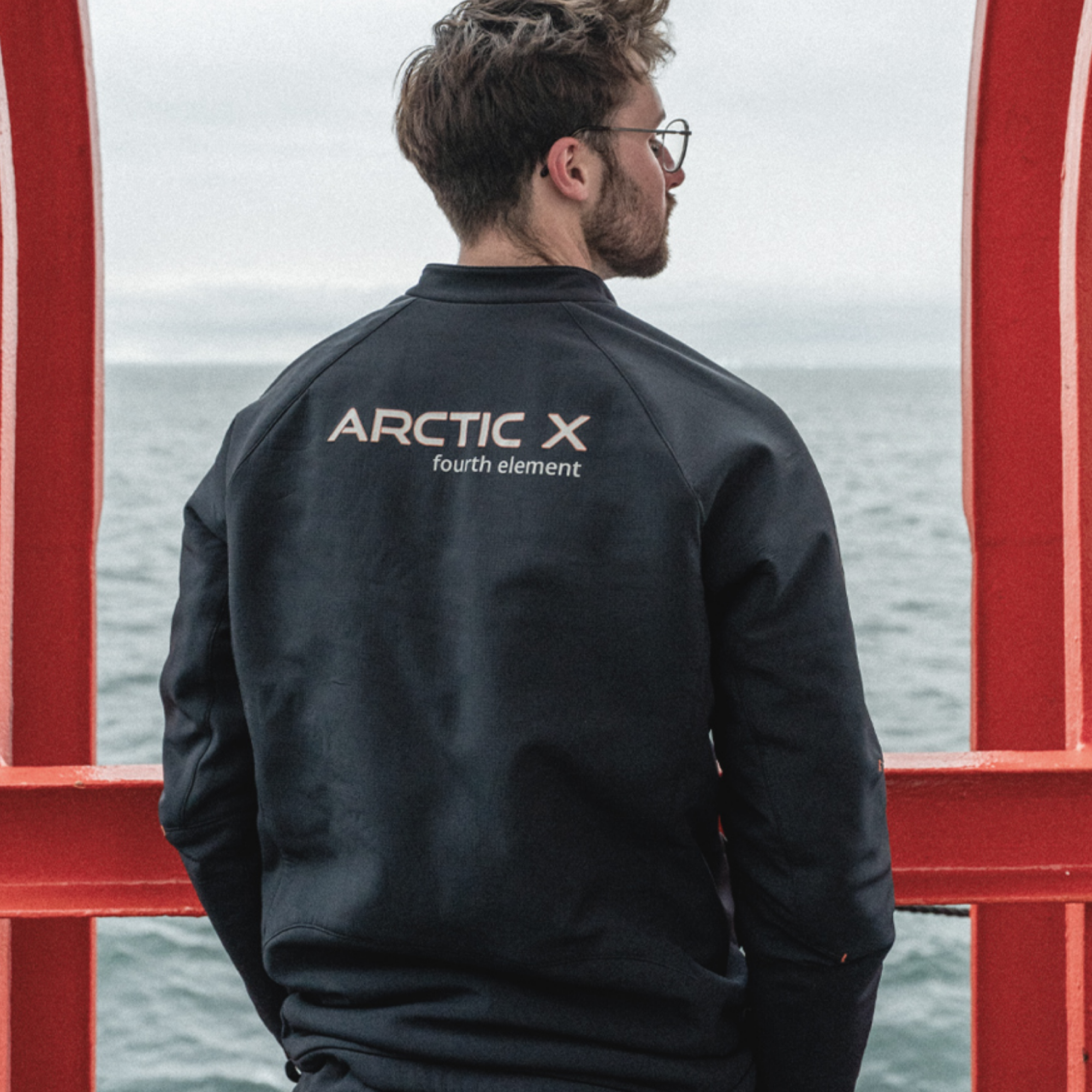 A man wearing a black jacket with 'ARCTIC X fourth element' printed on the back, standing in front of a red metal structure with an overcast sea background.