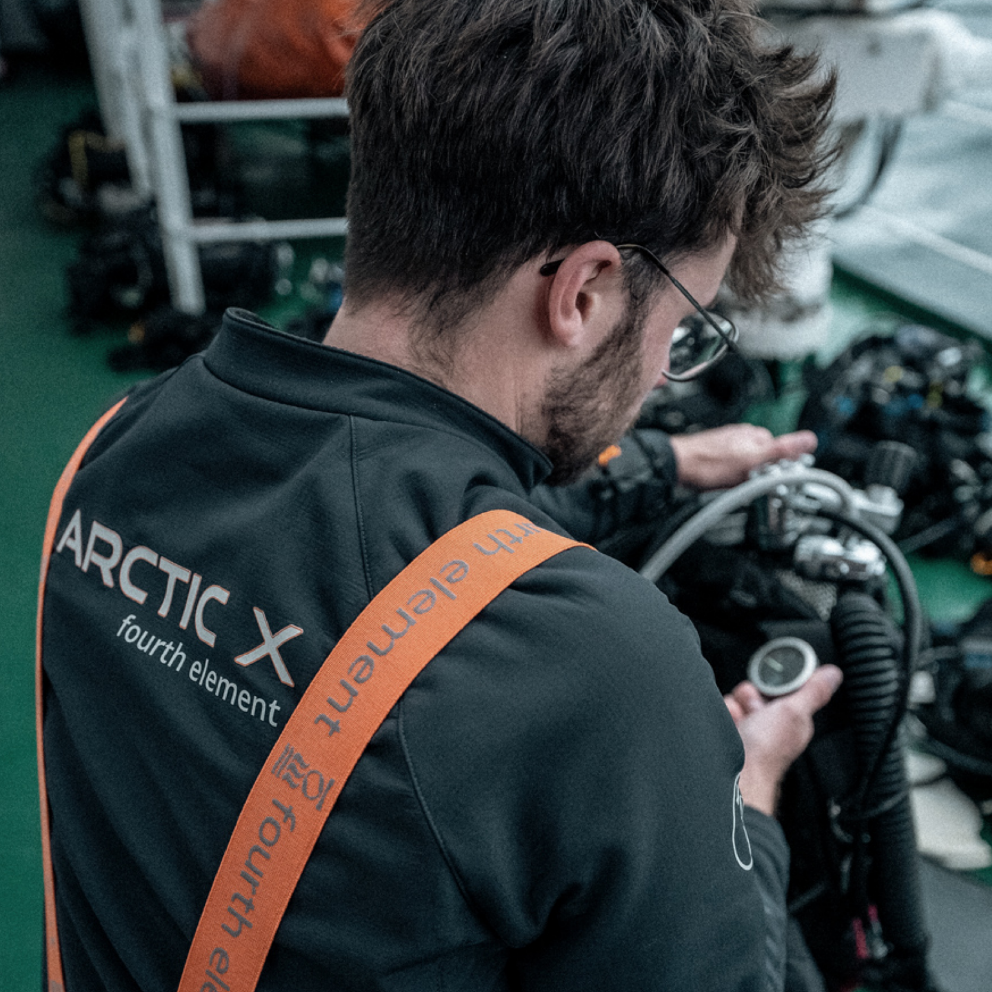 A man wearing a black Fourth Element Arctic X jacket with orange straps, seen from behind as he works with diving equipment in a workshop setting.