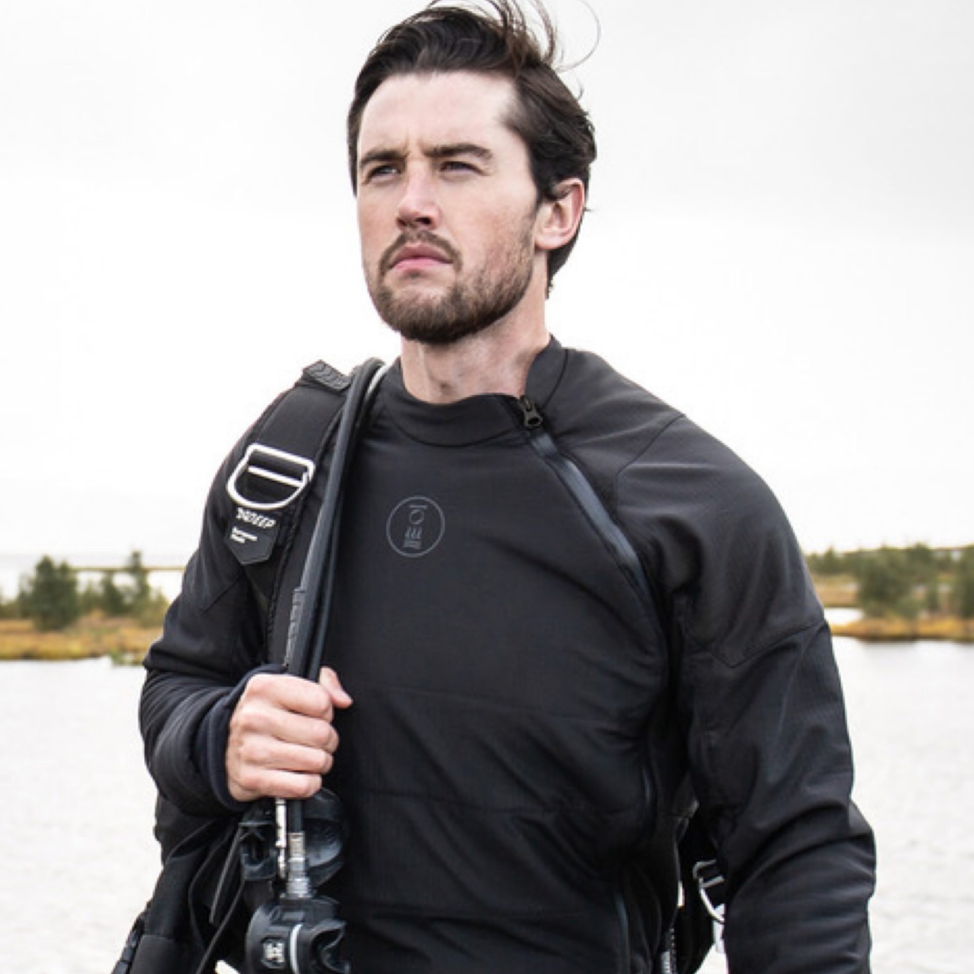 A man wearing a black technical wetsuit top and carrying a black diving backpack outdoors near water under an overcast sky.