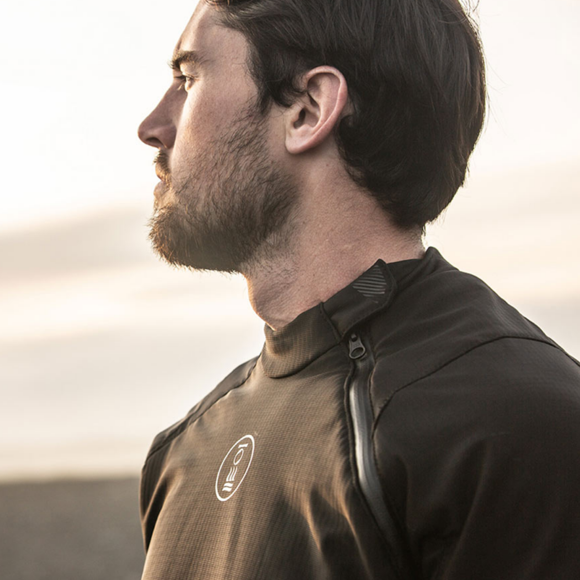 A man in profile wearing a dark brown technical outdoor jacket with a high collar and a zippered chest pocket, set against a soft, warm-toned blurred background.