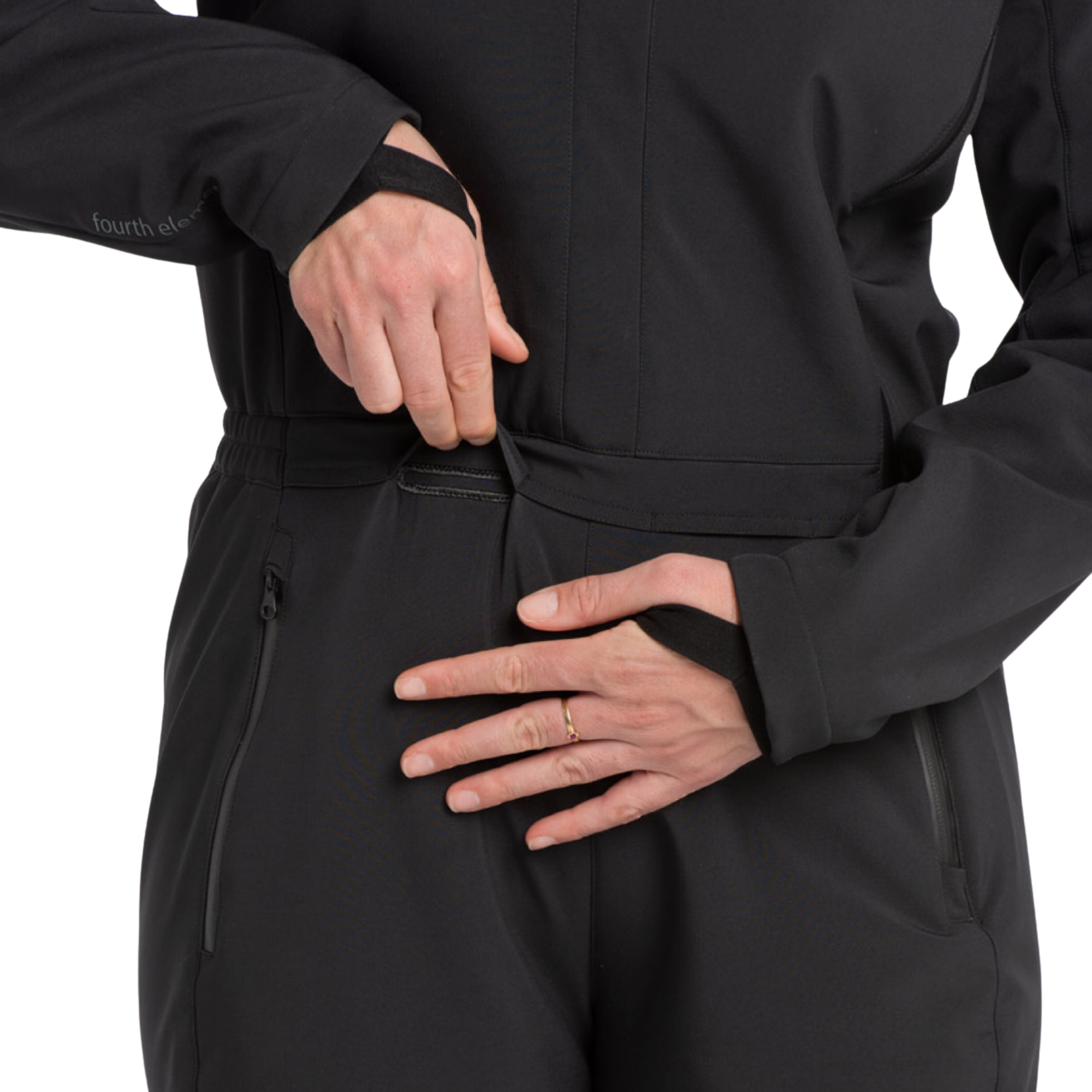 Close-up of a person wearing a black technical one-piece jacket or suit, showing the waist zipper and side zip pockets, with hands demonstrating fit and closure. The brand name 'fourth element' is subtly printed on the left sleeve.