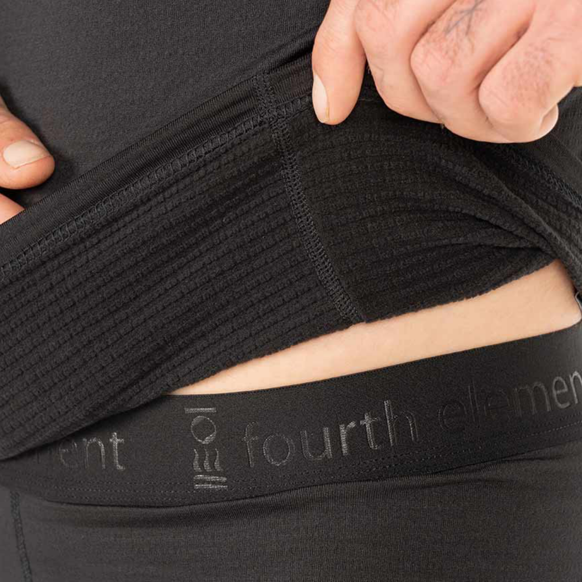 Close-up of a person lifting a black ribbed garment or undergarment to reveal the waistband, which has the text 'fourth element' and a small logo. The hands are visible pulling the fabric upward.