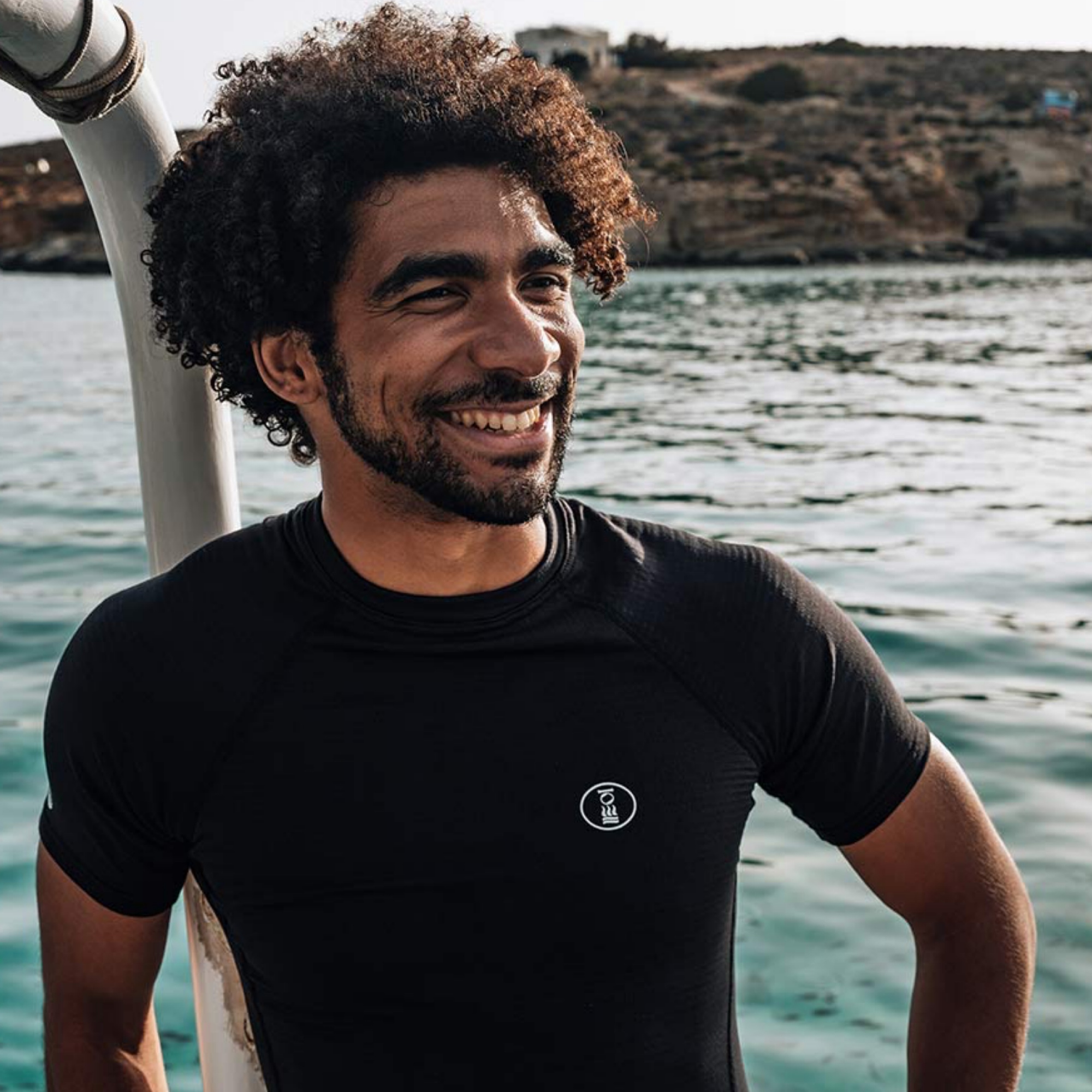 A smiling man with curly dark hair wearing a black short-sleeve rash guard swimsuit with a small white circular logo on the chest, standing on a boat with water and a rocky shoreline in the background.
