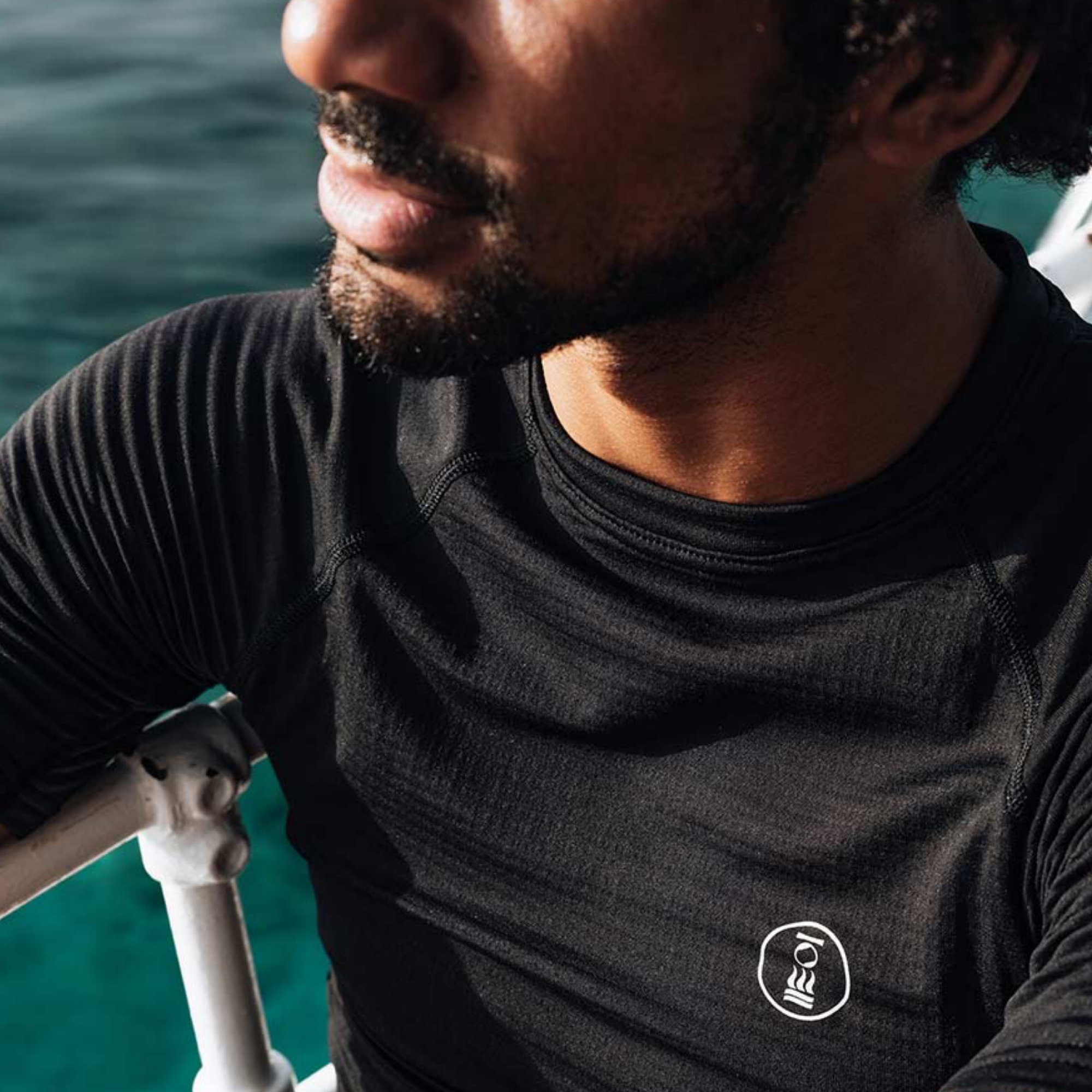 A close-up of a man wearing a black long-sleeve athletic shirt with a small white circular logo on the chest, set against a background of blue-green water.