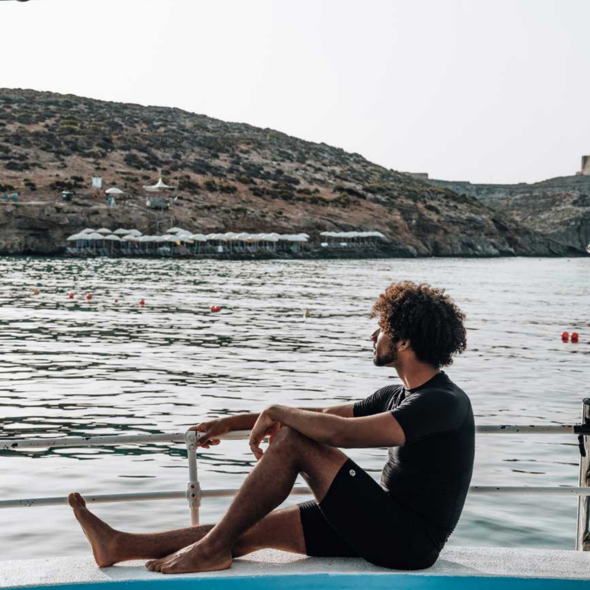 A man sits barefoot on the edge of a boat or dock, looking out over calm water toward a rocky coastal hillside with small buildings in the distance. He is wearing a black short-sleeve top and black shorts.
