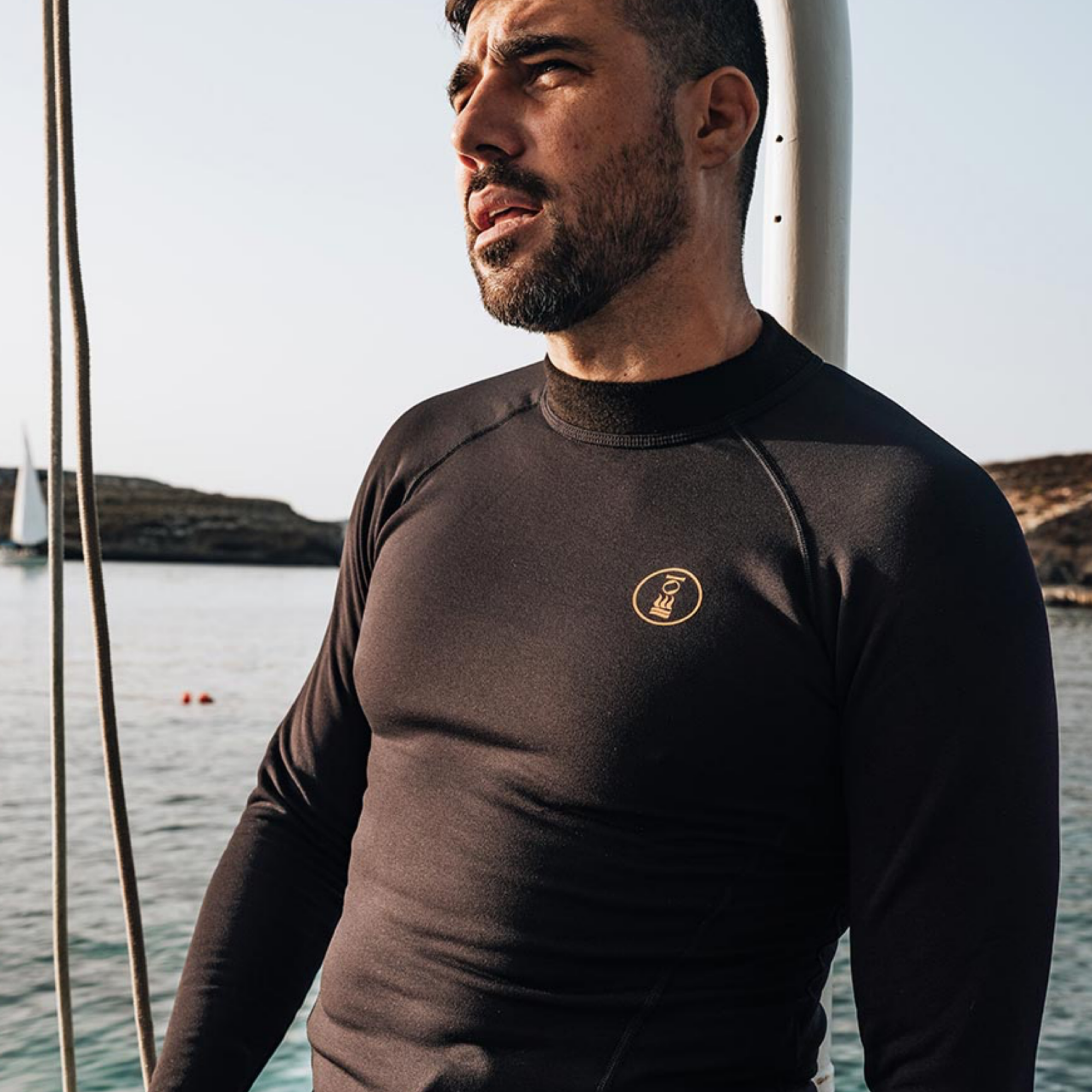 A man wearing a black long-sleeve wetsuit top with a gold circular logo on the chest, standing on a boat with water and distant land visible in the background.