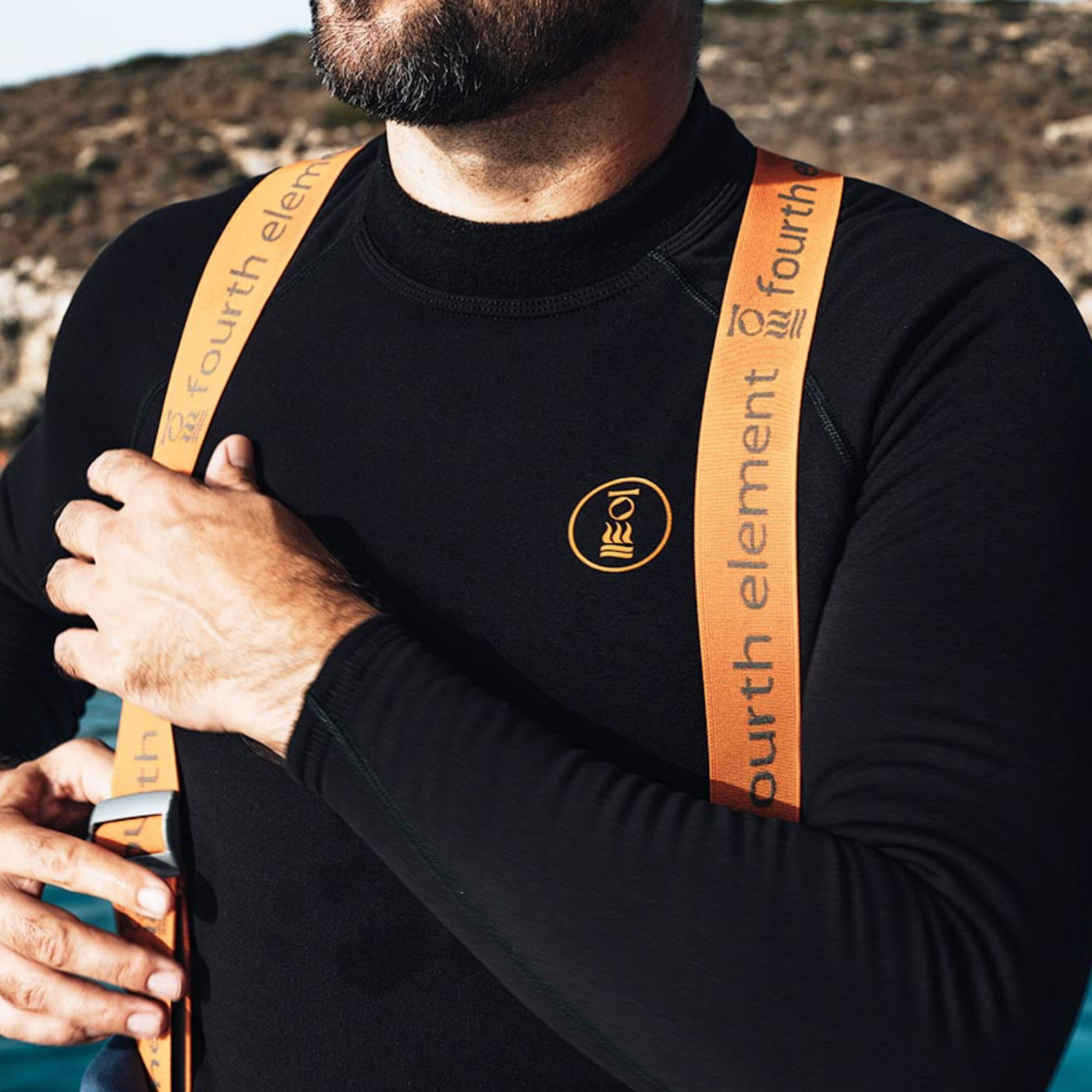 A close-up of a man wearing a black long-sleeve wetsuit top with orange shoulder straps and a small circular orange chest logo. The straps are printed with the brand name 'fourth element'.