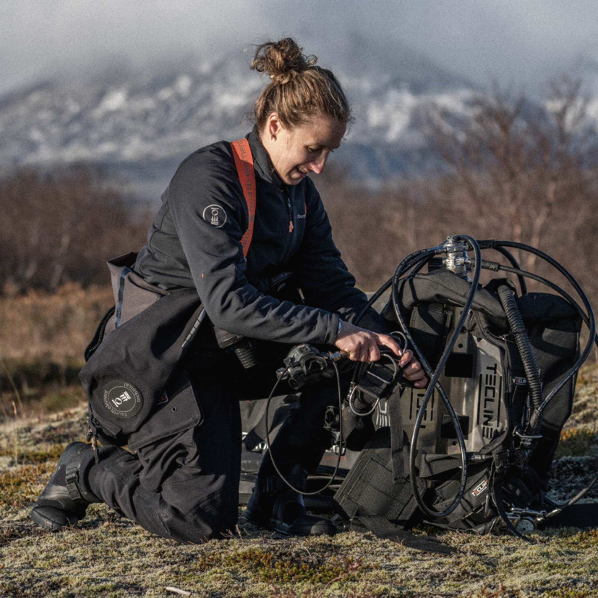 A woman kneels outdoors in a grassy field with mountains in the background, adjusting a large technical diving or scuba gear setup on the ground. She is wearing a dark waterproof jacket and pants, and the equipment is mostly dark gray and black with some orange accents.