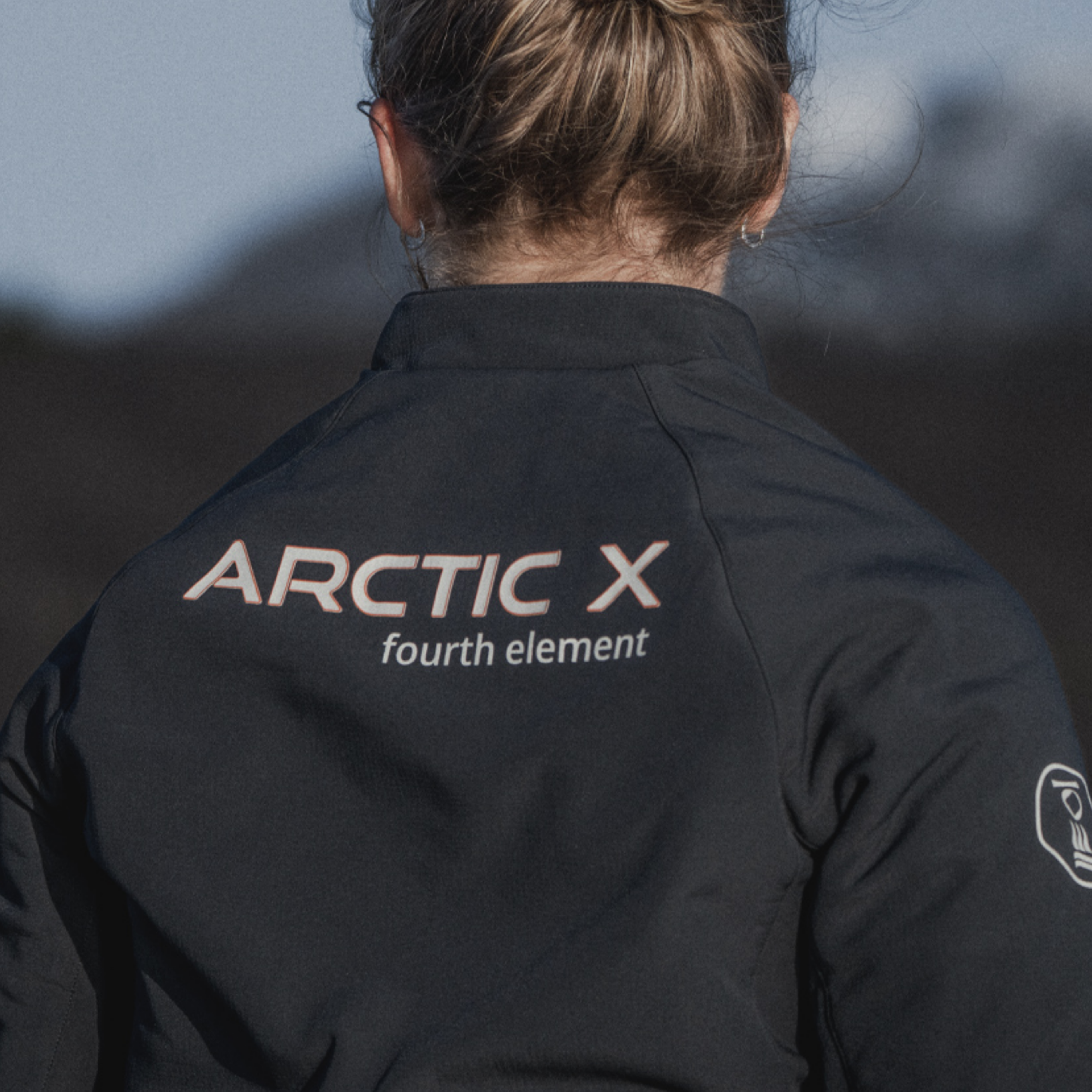 Back view of a person wearing a black Arctic X jacket by fourth element, with the brand name printed in white on the upper back, set against a blurred outdoor background.