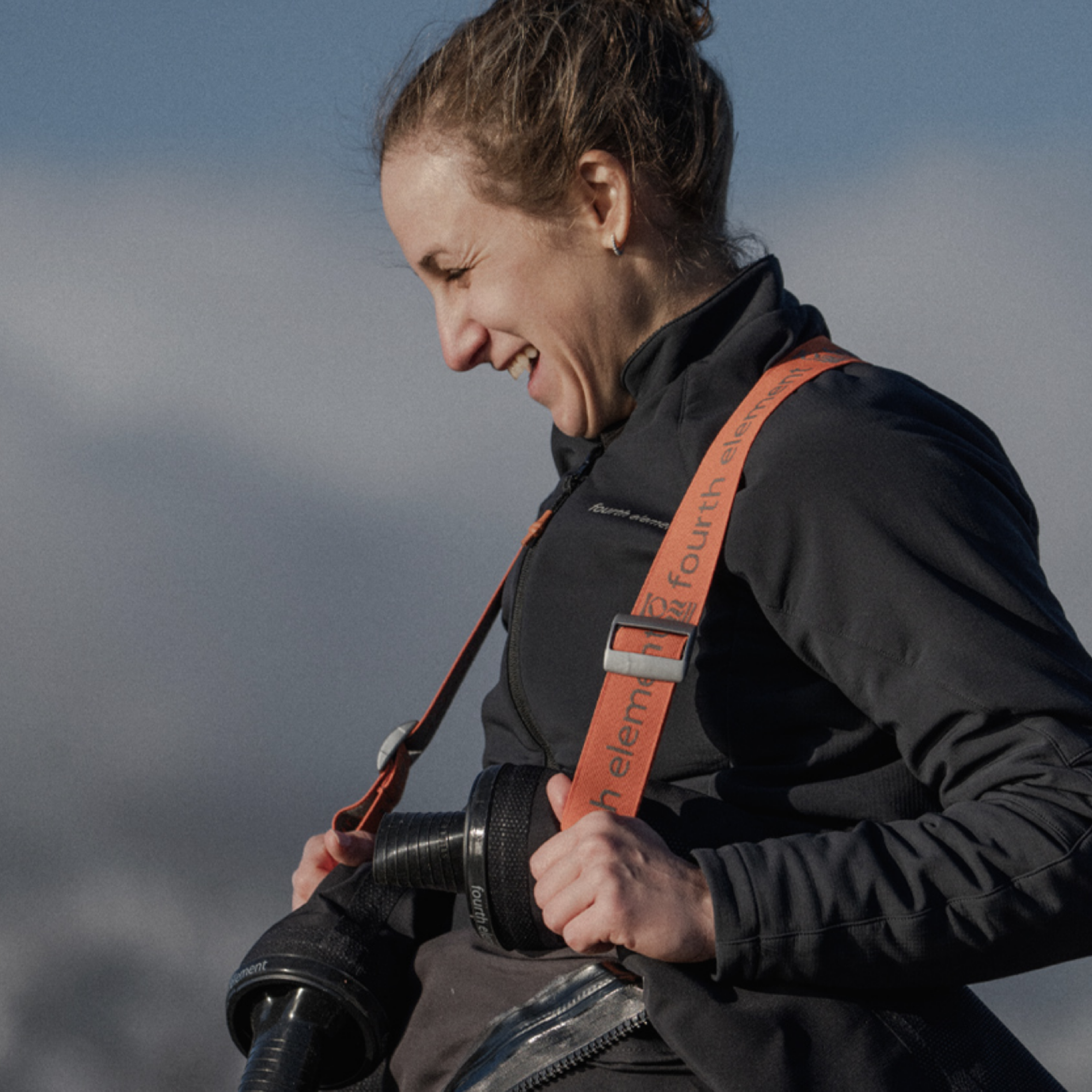 A woman outdoors wearing a black jacket and holding a camera with an orange shoulder strap across her chest.