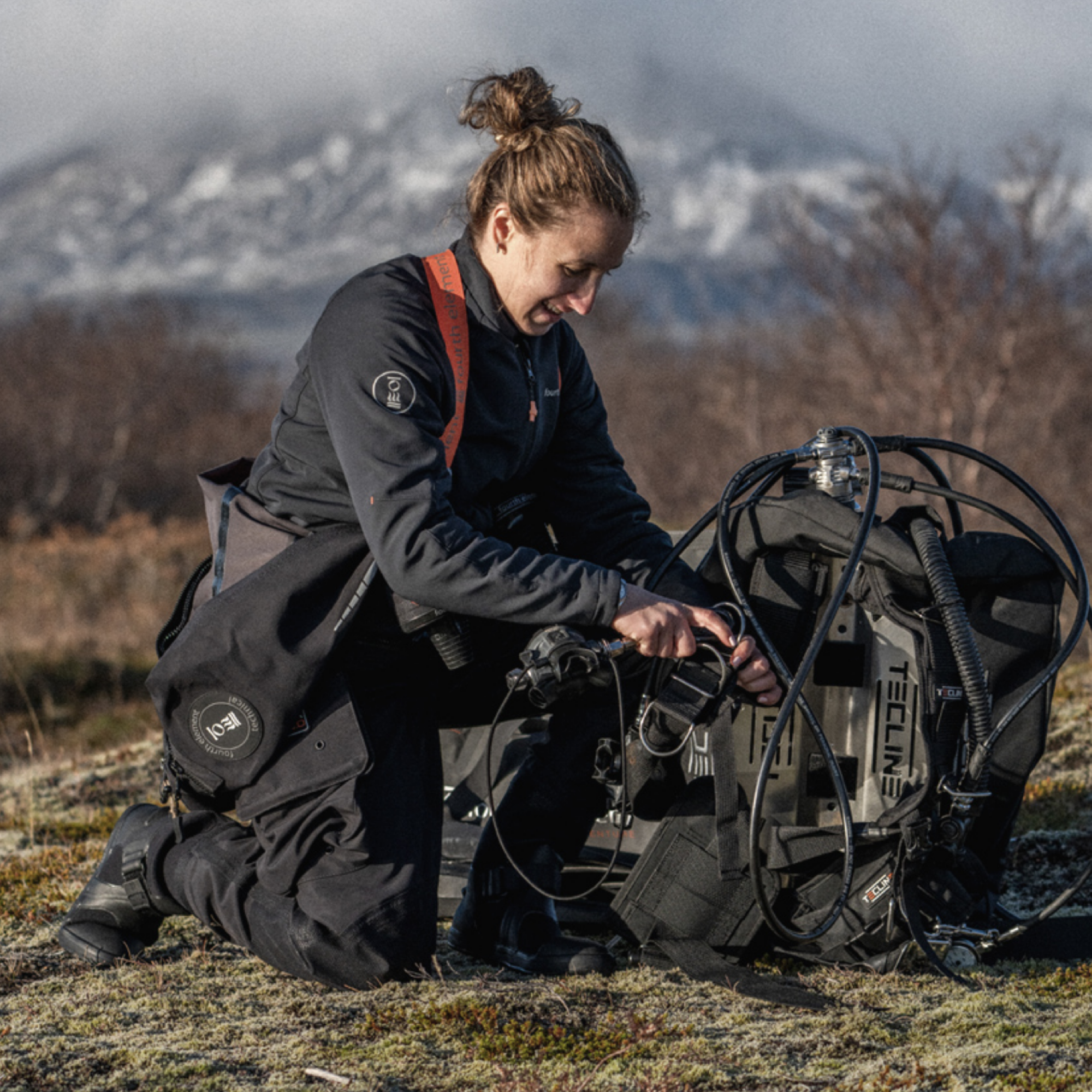 A woman in black outdoor gear kneels on grassy ground next to a large technical diving or submersible equipment rig, with snowy mountains in the background.
