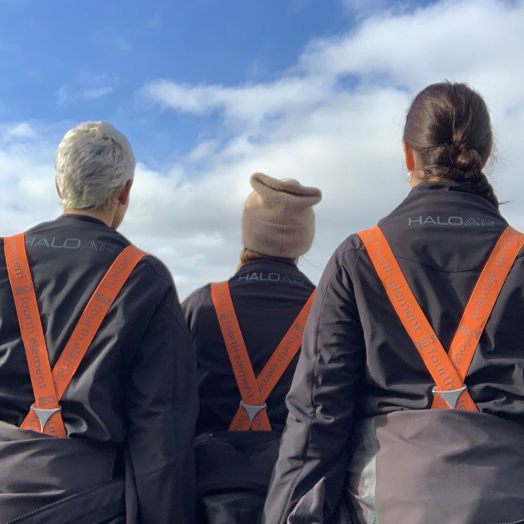 Three people seen from behind wearing matching dark HALO branded jackets with bright orange suspenders, standing outdoors against a blue sky with clouds.