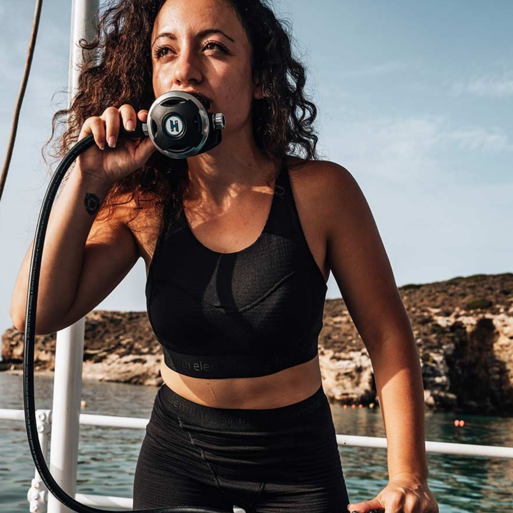 A woman on a boat wearing a black athletic sports bra and black leggings, holding a scuba mask near her face. The background shows water, rocky coastline, and blue sky.