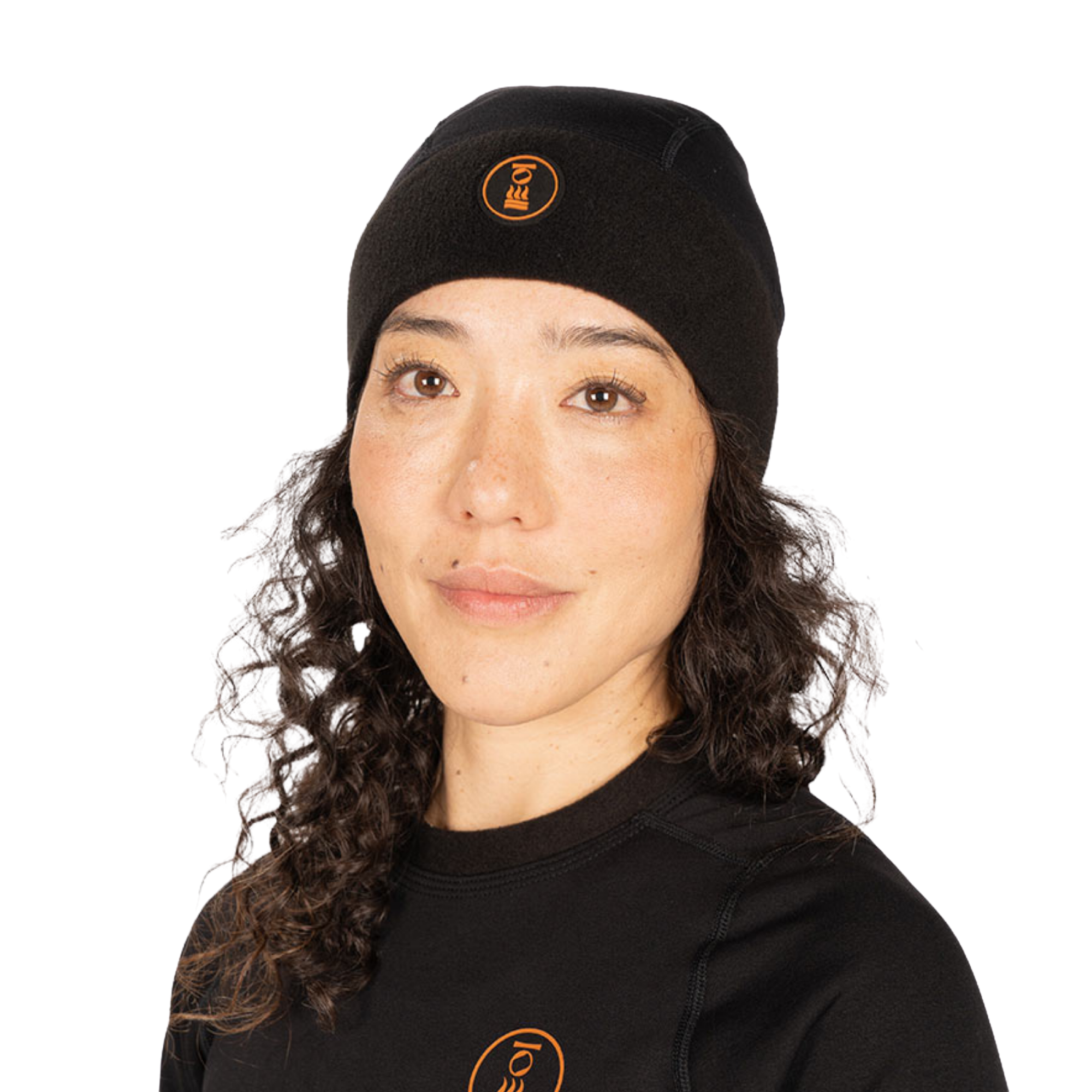A woman with long curly dark hair wearing a black knit beanie and a black long-sleeve top, both featuring a small circular orange logo on the front, photographed against a plain white background.