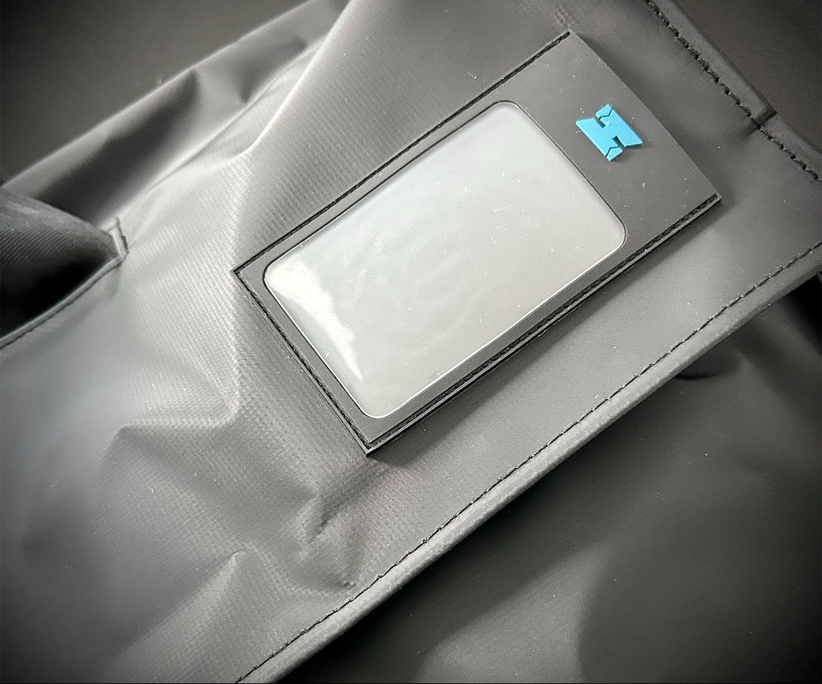 Close-up of a gray fabric sleeve or pouch with a rectangular clear plastic window and a small blue logo, shown on a dark gray background. Halcyon Traverse Bag for divers. 