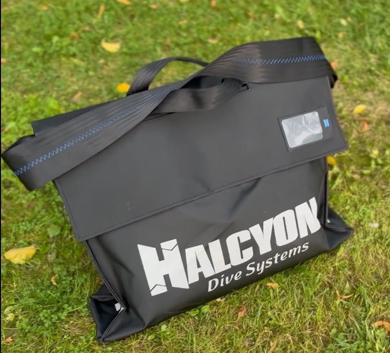 A black Halcyon Dive Systems Traverse Bag carrying bag with white logo text, resting on a green grassy outdoor lawn. Ideal for divers. 