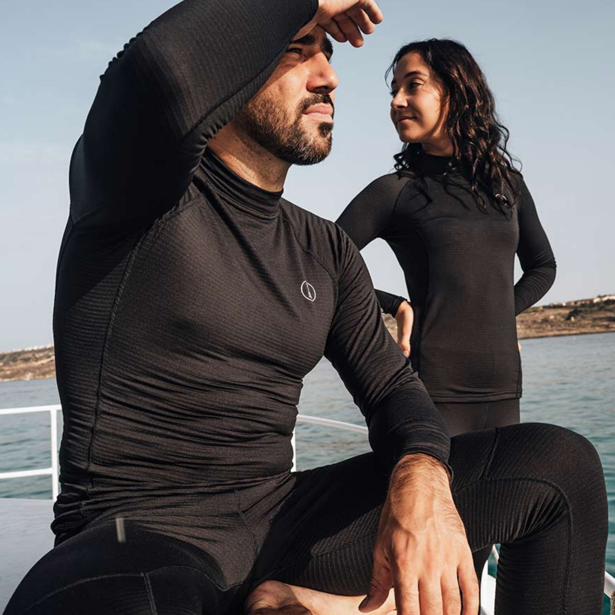 A man and a woman wearing matching black long-sleeve athletic wetsuits sit on a boat outdoors by the water.