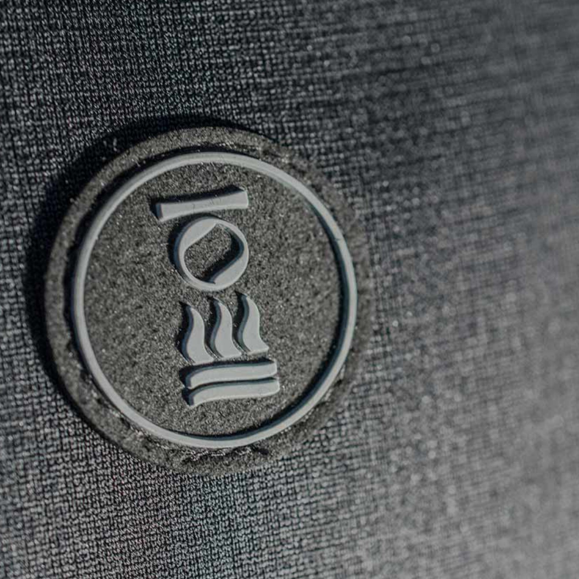 A close-up of a circular dark gray patch with a white stylized logo, mounted on a textured dark gray fabric background.