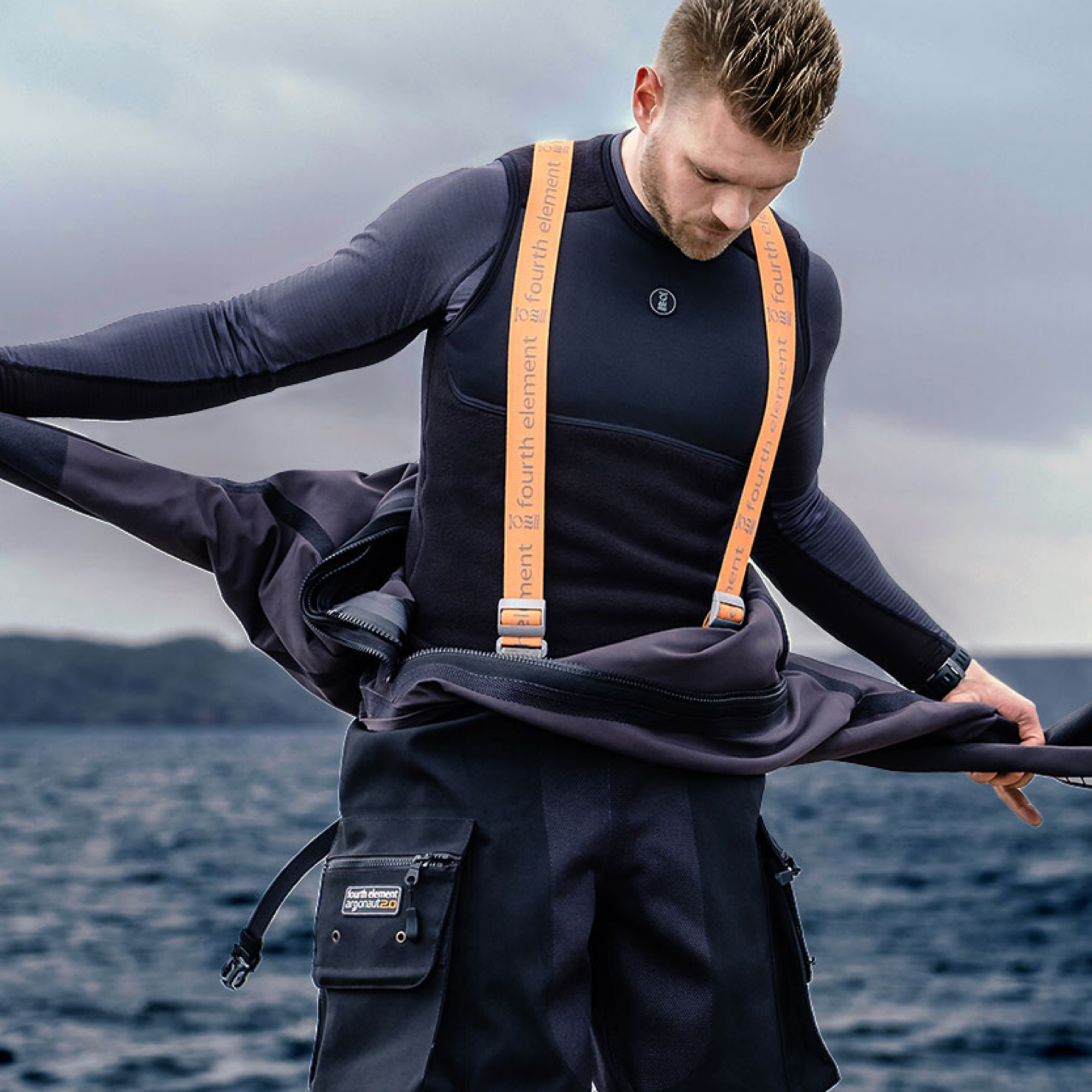 A man wearing a black wetsuit and orange suspenders stands by the ocean, holding open a black wet jacket. 