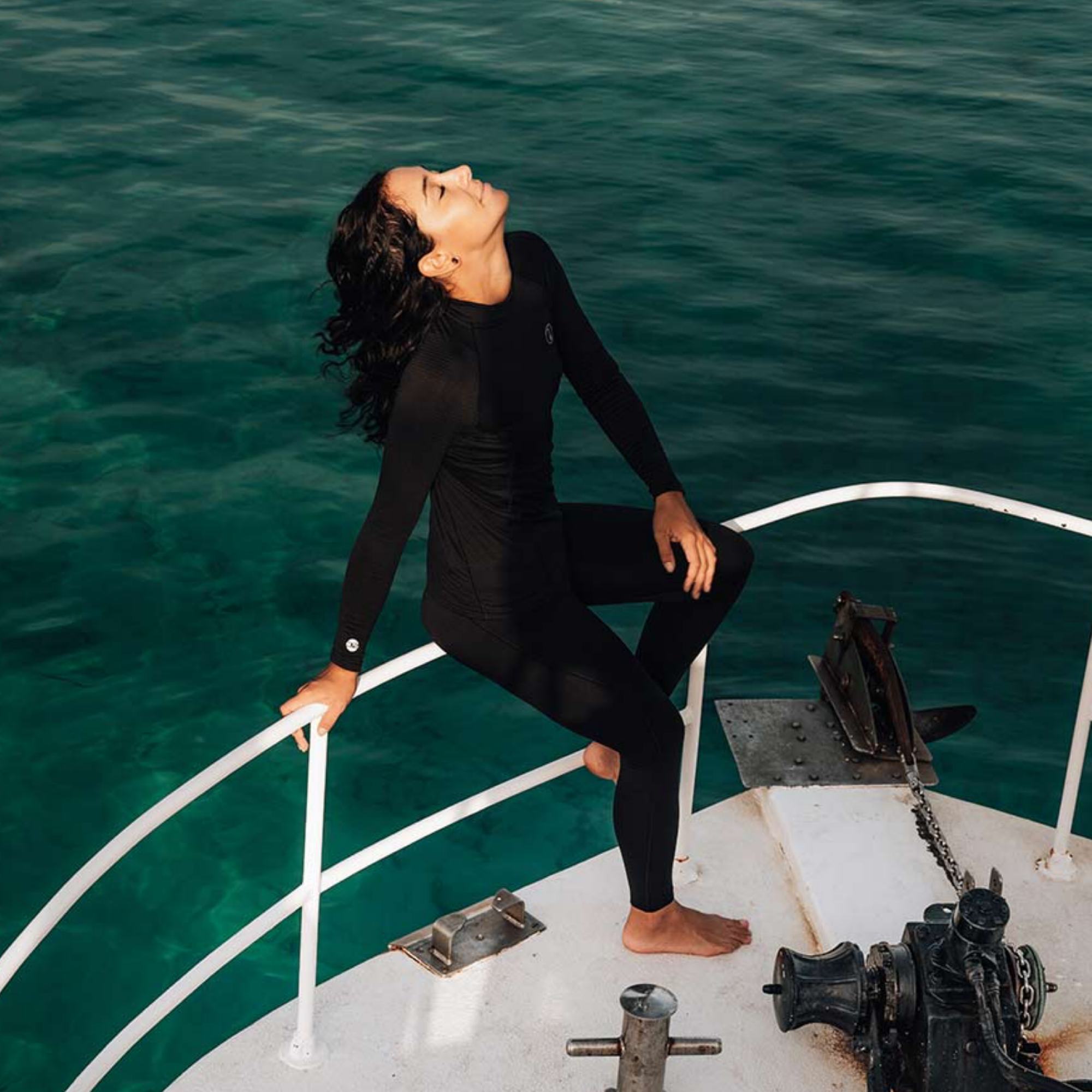 A woman in a black long-sleeve wetsuit sits barefoot on the white deck of a boat, leaning against the railing with her head tilted back, with deep green-blue ocean water in the background.