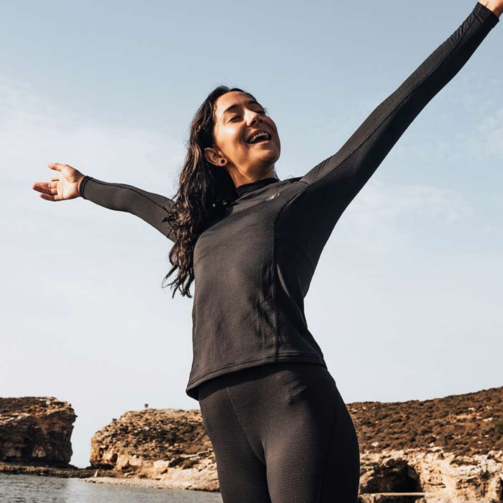 A woman with long dark hair stands outdoors with her arms outstretched, wearing a fitted black long-sleeve top and matching black leggings against a pale sky and rocky coastal background.