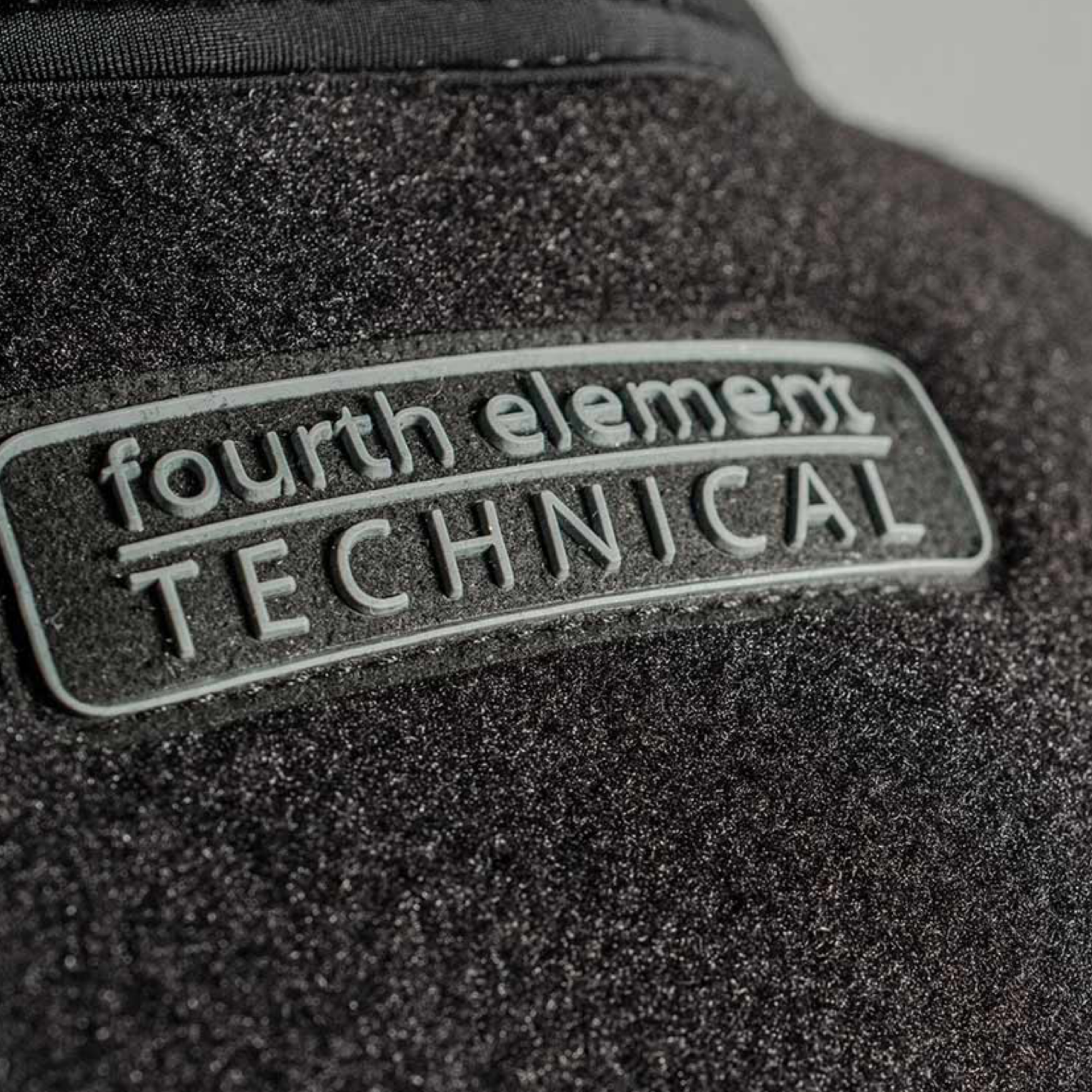 Close-up of a black textured technical fabric with a rectangular metallic silver patch reading 'fourth element TECHNICAL'.